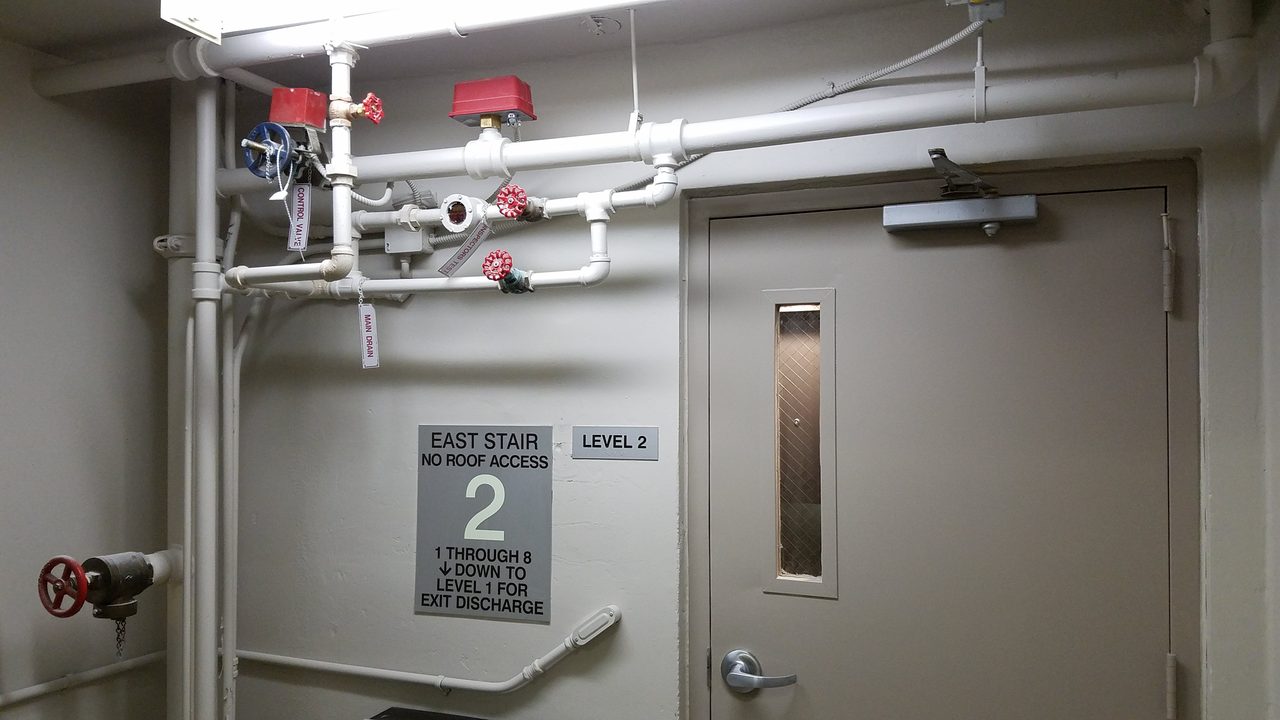 Hollow metal stairwell fire door with a closer and narrow vision panel, with East Stair and Level 2 signs mounted on the wall beside it